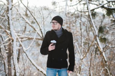 Bir gri kazak ve siyah ceket siyah bir şapka ve gözlük ile genç bir adam portresi kış parkta sıcak kahve içer