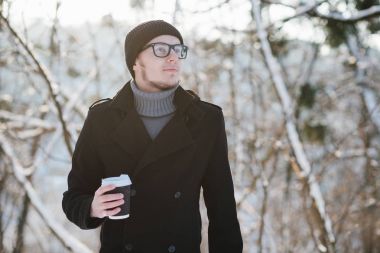 Bir gri kazak ve siyah ceket siyah bir şapka ve gözlük ile genç bir adam portresi kış parkta sıcak kahve içer