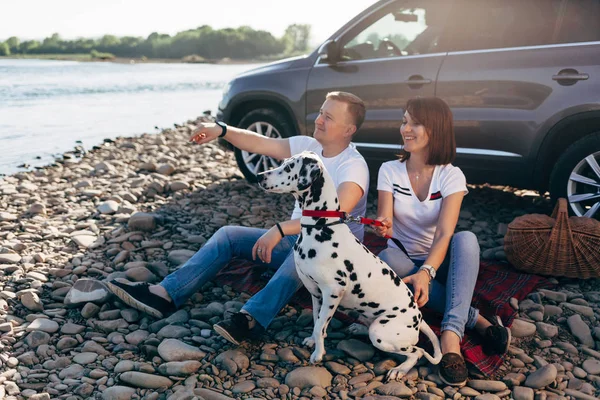幸福的年轻夫妇与狗在 roadtrip 的肖像。男人和女人坐在格子上户外野餐概念. — 图库照片