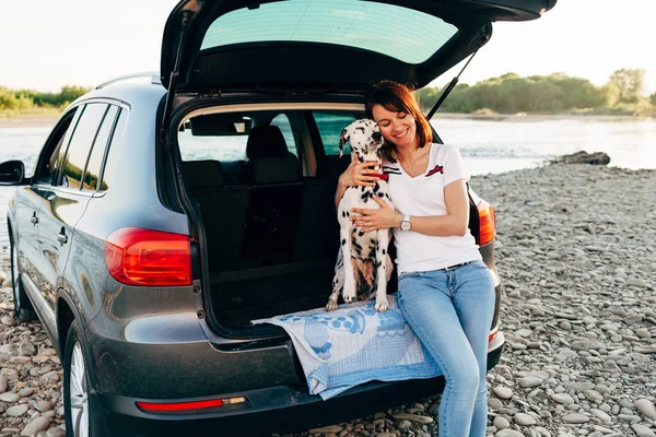 Portrait de femme joyeuse assise en voiture avec chien regardant le coucher du soleil par la fenêtre, profiter du concept de voyage . — Photo