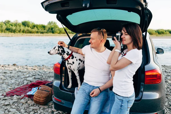 Portrait de couple heureux de jeunes adultes avec chien en voyage. Homme assis sur plaid avec une femme. Concept de pique-nique extérieur . — Photo