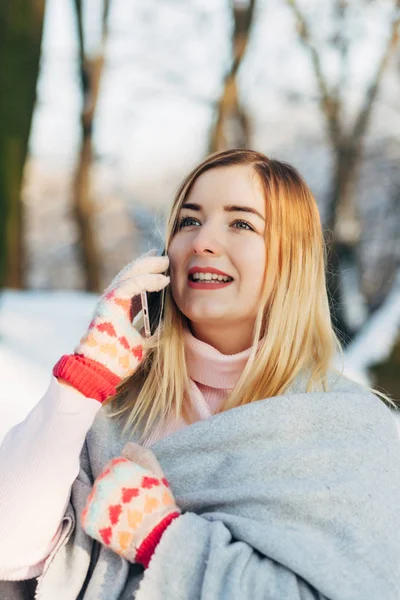 ピンクのセーターとニットのスカーフで若い白人魅力的なブロンドの女の子が冬の公園でのスマート フォンを屋外の保持します。 — ストック写真