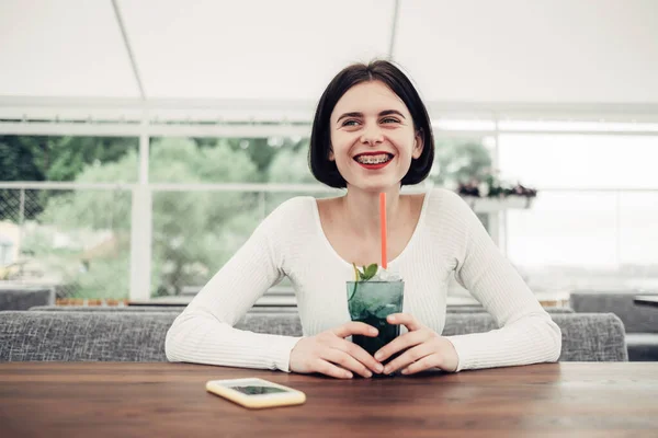 Primer plano retrato de hermosa morena chica en suéter blanco sonrisa feliz con ortodoncia bebidas cóctel —  Fotos de Stock