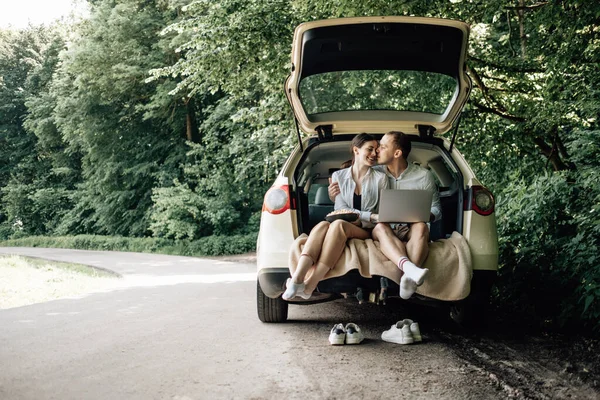 Jovem casal feliz vestido de igual em T-shirt branca Sentado no tronco do carro com laptop e pipoca na beira da estrada, fim de semana fora da cidade, feriados e Road Trip Concept — Fotografia de Stock