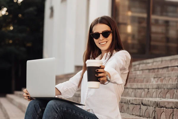 Retrato de uma menina elegante vestida em jeans e camisa branca segurando laptop e café quente, trabalhador freelance, senhora de negócios, conceito de poder da mulher — Fotografia de Stock