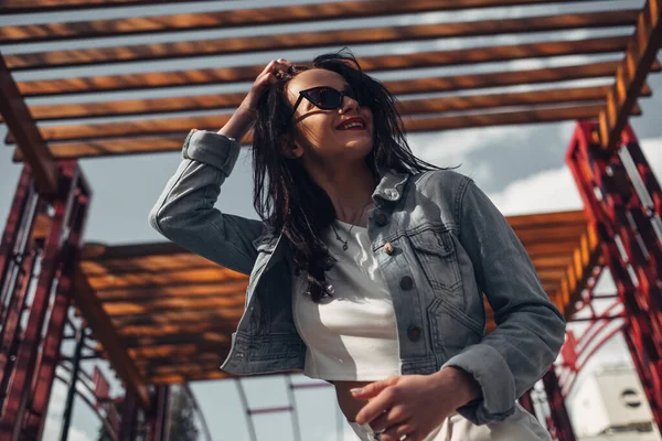 Schöne brünette Mädchen mit Sonnenbrille, weißer Hose und Jeansjacke, rotem Lippenstift — Stockfoto