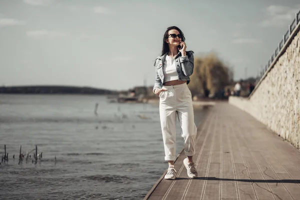 Belle fille brune en lunettes de soleil pantalon blanc et veste de jeans près du lac — Photo