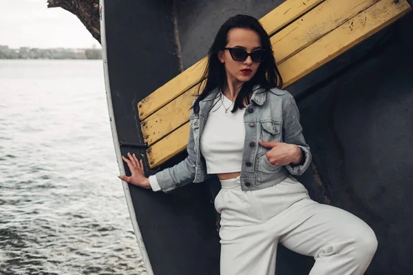 Hermosa chica morena en gafas de sol pantalones blancos y Jeans chaqueta cerca del lago —  Fotos de Stock