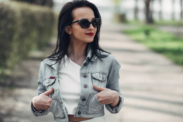 Vacker brunett flicka i solglasögon vita byxor och jeans jacka, rött läppstift — Stockfoto