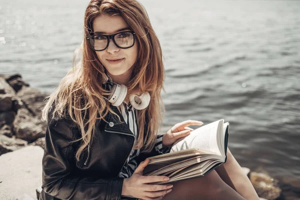 Retrato de verão ao ar livre da menina bonita nova em jaqueta de couro preto e óculos lê o livro perto do lago — Fotografia de Stock