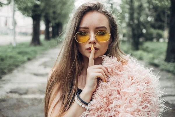 Portret van een stijlvolle meisje in roze vacht en moderne gele zonnebril poseren op straat — Stockfoto