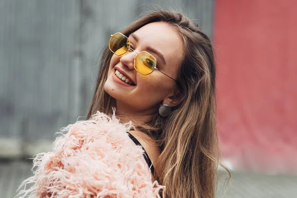 Portret van een stijlvolle meisje in roze vacht en moderne gele zonnebril poseren op straat — Stockfoto