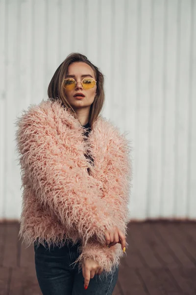 Porträt eines stilvollen Mädchens in rosa Mantel und moderner gelber Sonnenbrille, das auf der Straße posiert — Stockfoto