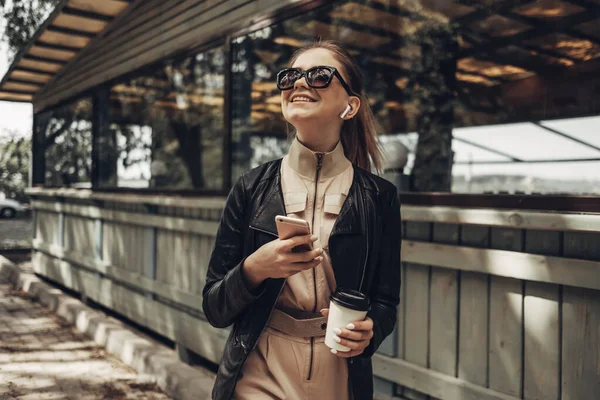 Портрет стильной девушки носит женский комбинезон и черный кожаный пиджак пьет кофе, используя смартфон — стоковое фото