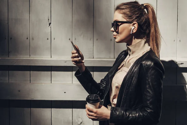 Retrato de elegante menina usa macacões mulheres e jaqueta de couro preto beber café, usando Smartphone — Fotografia de Stock