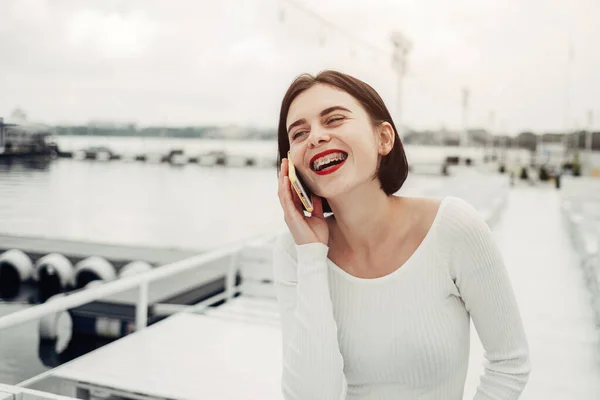 Портрет красивой брюнетки в белом свитере счастливая улыбка с зубными брекетами разговаривая по мобильному телефону — стоковое фото