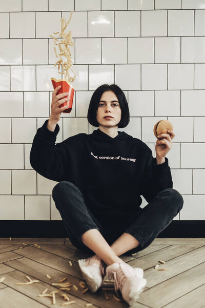Beautiful Brunette Girl in Black Pullover with Text ''Best Version of Yourself" Sitting on the Floor Throws French Fries Stock Photo