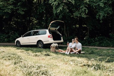 Beyaz tişörtlü genç mutlu çift arabanın yanında eğleniyor, hafta sonu şehir dışında, tatiller ve yolculuk konsepti.