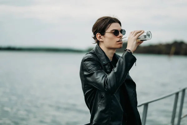 Close Up Portret van een stijlvolle jongen met zonnebril gekleed in lederen jas en zwart T-shirt Drinken Bier op de pier in de buurt van de rivier — Stockfoto