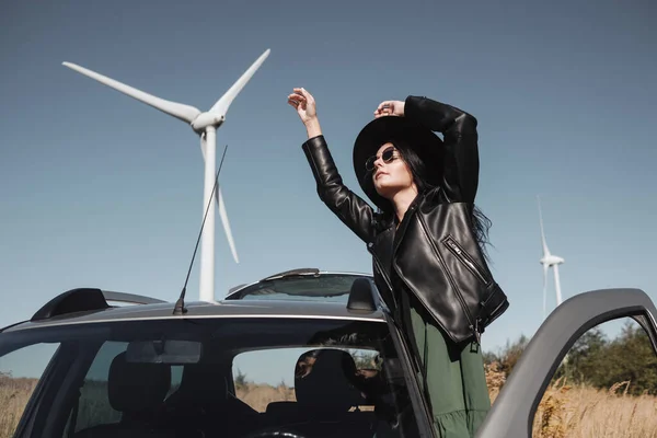 Happy Traveling Girl Enjoying a Car Trip on the Field Road with Electric Wind Turbine Power Generator on the Background