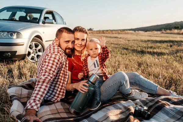 Szczęśliwa młoda rodzina Mama i tata z ich małym synem korzystających letni weekend Piknik siedzi na kratę w pobliżu samochodu poza miastem w polu w słoneczny dzień Zachód słońca, wakacje i wycieczka koncepcja — Zdjęcie stockowe