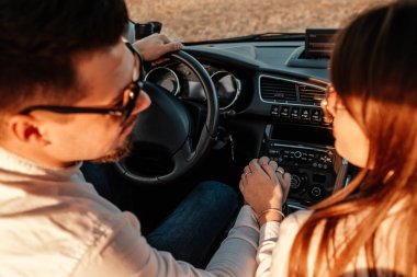Beyaz gömlekli ve kot pantolonlu genç mutlu çift yeni arabalarında yolculuğun tadını çıkarıyor, Sahada güzel bir gün batımı, tatil ve seyahat konsepti
