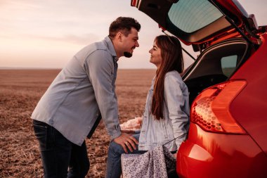 Beyaz Gömlekli ve Kot Giyen Genç Mutlu Çift Yeni Araba Bagajlarında Oturuyor, Sahada Güzel Günbatımı, Tatil ve Seyahat Konsepti