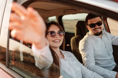 Beyaz gömlekli ve kot pantolonlu genç mutlu çift yeni arabalarında yolculuğun tadını çıkarıyor, Sahada güzel bir gün batımı, tatil ve seyahat konsepti