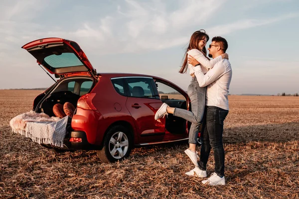 Joven pareja feliz vestida como en camisa blanca y pantalones vaqueros disfrutando de un viaje por carretera en su nuevo coche, hermoso atardecer en el campo, vacaciones y concepto de viaje —  Fotos de Stock