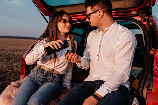 Young Happy Couple Dressed Alike in White Shirt and Jeans Enjoying Road Trip at Their New Car, Beautiful Sunset on the Field, Vacation and Travel Concept — 스톡 사진