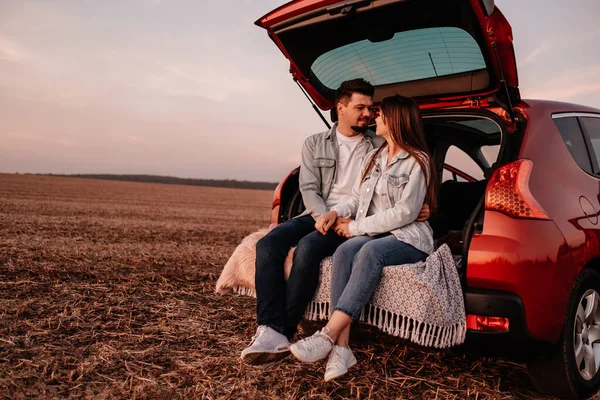 Young Happy Couple Dressed Alike in White Shirt and Jeans Sitting at Their New Car Trunk, Beautiful Sunset on the Field, Vacation and Travel Concept — Stock Photo, Image