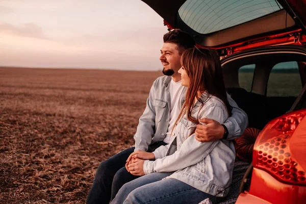 Joven pareja feliz vestida como en camisa blanca y pantalones vaqueros sentados en su nuevo baúl de coche, hermoso atardecer en el campo, vacaciones y concepto de viaje — Foto de Stock