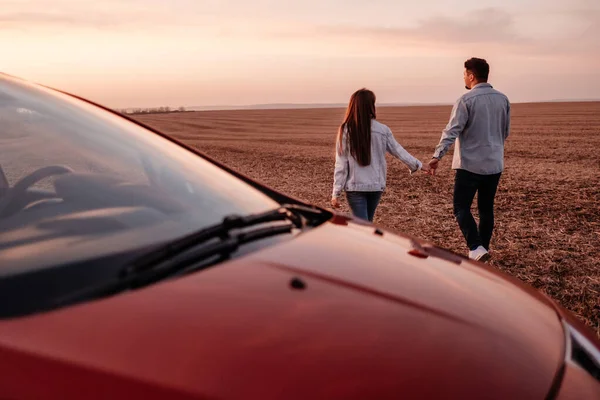 Jovem casal feliz vestido de igual em camisa branca e jeans desfrutando de viagem rodoviária em seu novo carro, belo pôr do sol no campo, férias e conceito de viagem — Fotografia de Stock