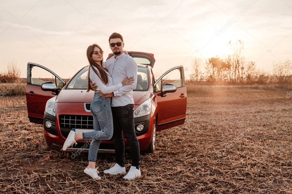 Young Happy Couple Dressed Alike in White Shirt and Jeans Enjoying Road Trip at Their New Car, Beautiful Sunset on the Field, Vacation and Travel Concept