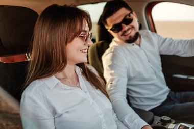 Beyaz gömlekli ve kot pantolonlu genç mutlu çift yeni arabalarında yolculuğun tadını çıkarıyor, Sahada güzel bir gün batımı, tatil ve seyahat konsepti