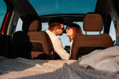 Beyaz gömlekli ve kot pantolonlu genç mutlu çift yeni arabalarında yolculuğun tadını çıkarıyor, Sahada güzel bir gün batımı, tatil ve seyahat konsepti