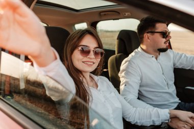 Beyaz gömlekli ve kot pantolonlu genç mutlu çift yeni arabalarında yolculuğun tadını çıkarıyor, Sahada güzel bir gün batımı, tatil ve seyahat konsepti