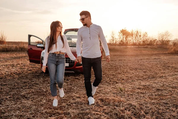Beyaz gömlekli ve kot pantolonlu genç mutlu çift yeni arabalarında yolculuğun tadını çıkarıyor, Sahada güzel bir gün batımı, tatil ve seyahat konsepti — Stok fotoğraf