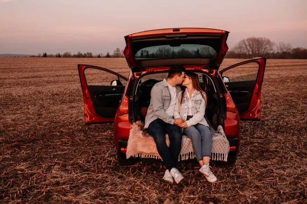 Jeune couple heureux habillé comme dans une chemise blanche et jeans assis à leur nouveau coffre de voiture, beau coucher de soleil sur le terrain, vacances et Voyage Concept — Photo