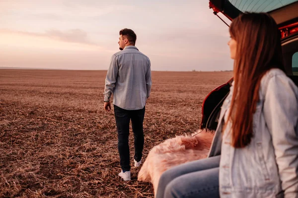 Молодые счастливые пара одеты как в белую рубашку и джинсы, сидя у своего нового автомобиля ствол, красивый закат на поле, отпуск и концепция путешествия — стоковое фото