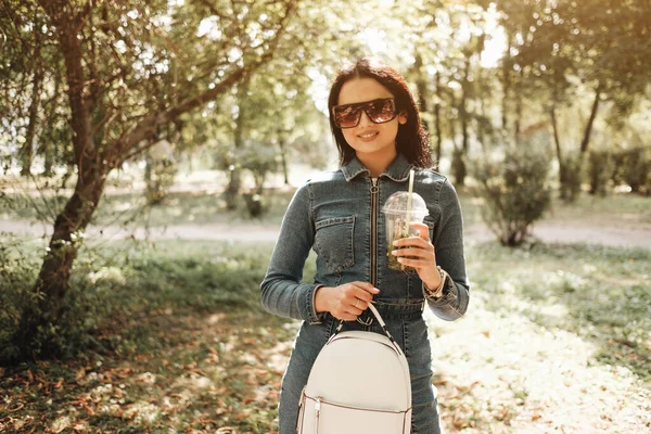 Retrato de uma bela morena menina vestida de jeans Denim macacão caminhadas no parque de outono — Fotografia de Stock