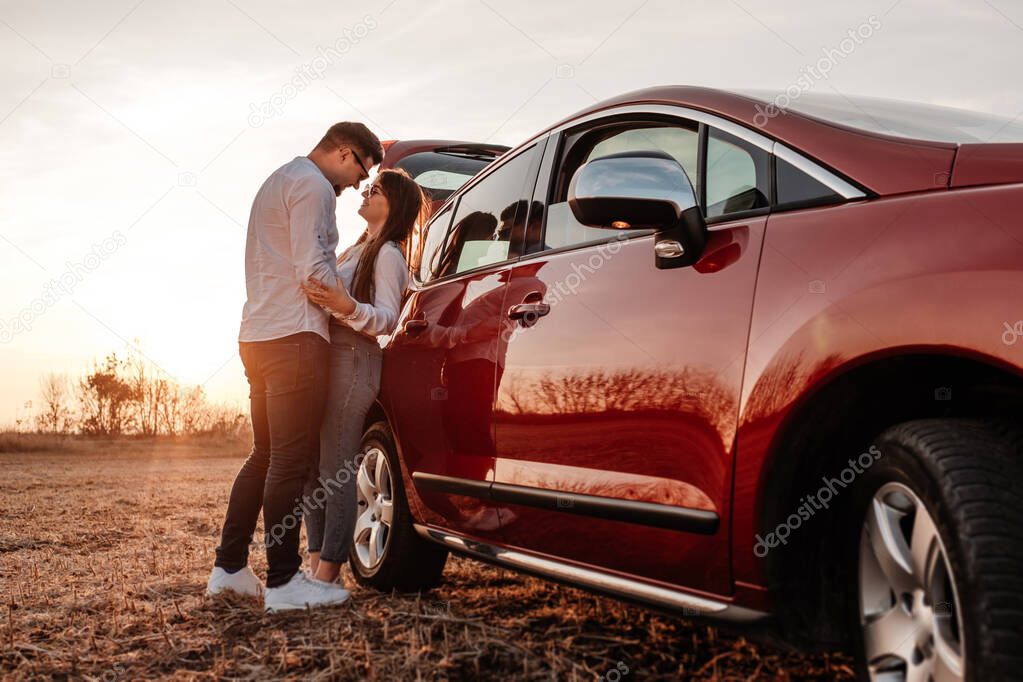 Young Happy Couple Dressed Alike in White Shirt and Jeans Enjoying Road Trip at Their New Car, Beautiful Sunset on the Field, Vacation and Travel Concept