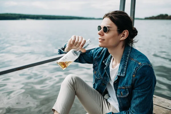 Retrato de cerca de un chico con estilo con gafas de sol vestidos con chaqueta vaqueros y camiseta blanca bebiendo cerveza en el muelle cerca del río — Foto de Stock