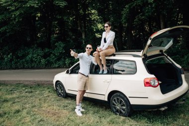 Beyaz tişörtlü genç mutlu çift arabanın yanında eğleniyor, hafta sonu şehir dışında, tatiller ve yolculuk konsepti.