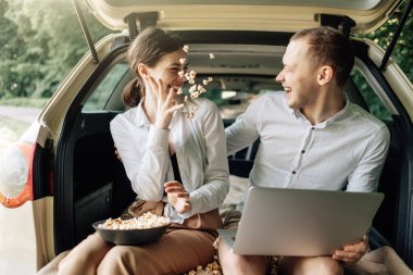 Genç, mutlu, beyaz tişörtlü bir çift. Araba bagajında dizüstü bilgisayar ve patlamış mısırla oturuyor. Hafta sonu şehrin dışında, tatillerde ve yolculukta.