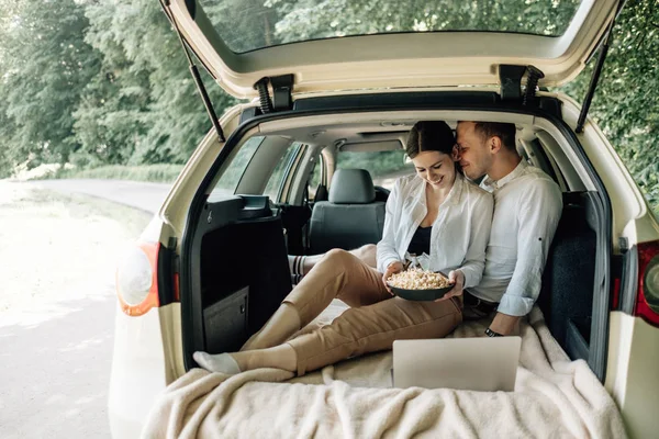 Joven Pareja Feliz Vestida Como en Camiseta Blanca Sentada en el Maletero del Coche con Portátil y Palomitas de Maíz en la Carretera, Fin de Semana Fuera de la Ciudad, Vacaciones y Concepto de Viaje por Carretera — Foto de Stock