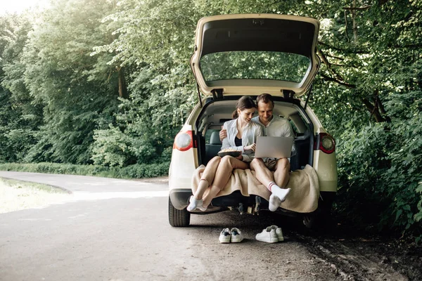 Joven Pareja Feliz Vestida Como en Camiseta Blanca Sentada en el Maletero del Coche con Portátil y Palomitas de Maíz en la Carretera, Fin de Semana Fuera de la Ciudad, Vacaciones y Concepto de Viaje por Carretera — Foto de Stock