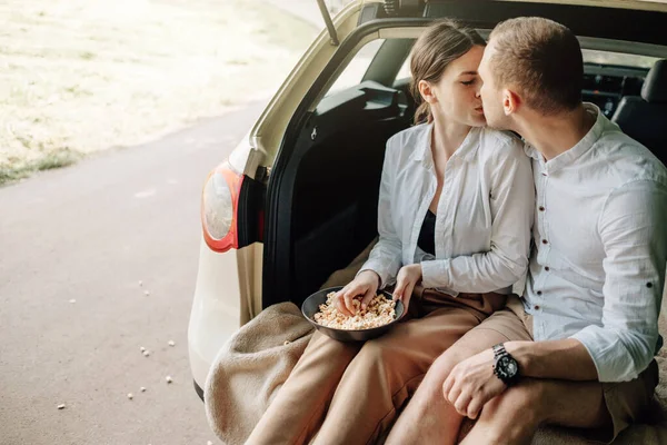 Fiatal boldog pár öltözött Alike fehér póló Ül az autó csomagtartóban popcorn az úton, hétvégén kívül a város, Ünnepek és Road Trip koncepció — Stock Fotó