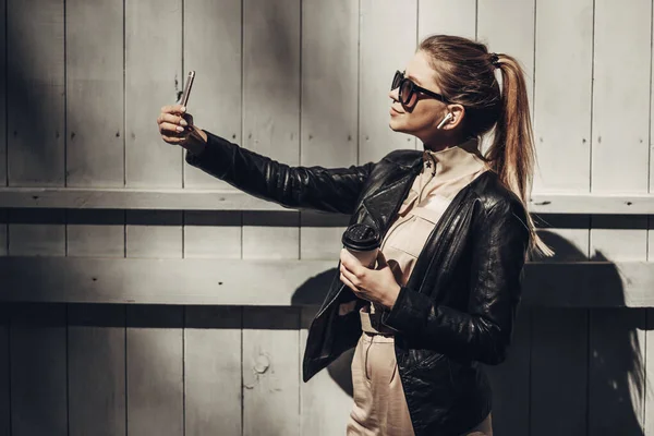 Portret stylowej dziewczyny nosi kobiet kombinezon i czarna skórzana kurtka pić kawę, korzystanie Smartphone — Zdjęcie stockowe