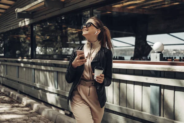 Πορτρέτο του κομψό κορίτσι φοράει γυναικεία φόρμα και μαύρο δερμάτινο μπουφάν ποτό καφέ, χρησιμοποιώντας smartphone — Φωτογραφία Αρχείου